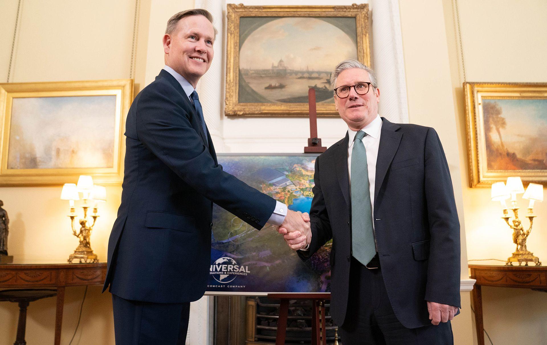 Prime Minister Sir Keir Starmer is shaking hands with the President of the Comcast Corporation, Mike Cavanagh, in front of a drawing of the proposed Universal park in a room in Downing Street. Either side of the men are lamps with three lightbulbs and lig